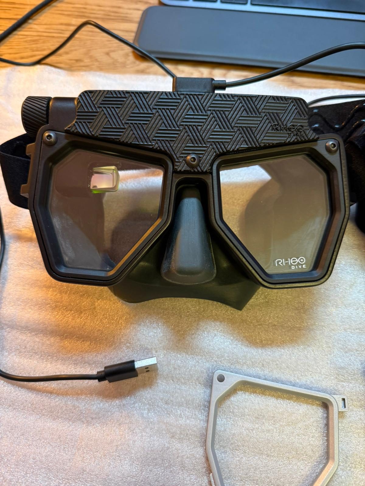 Rheo Dive mask on engineering desk with USB cable connected and CNC machined frame part — HUD optics visible
